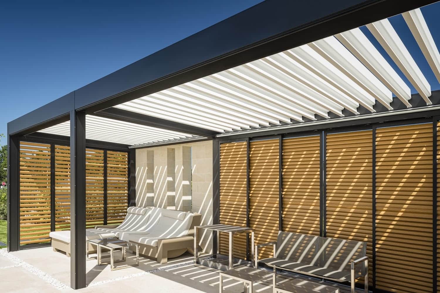 Sammenbygget Renson Camargue pergola med mørkt stel og lyse lameller. Siderne er monteret med Loggia skodder